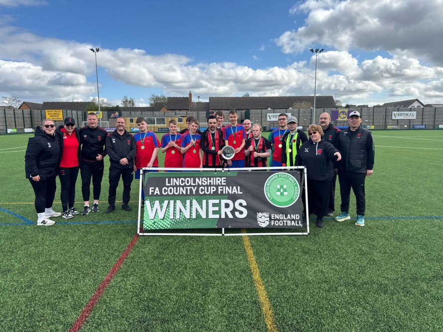 A group of people in football gear stood on a pitch. The banner in front of them says Lincolnshire FA County Cup Final Winners