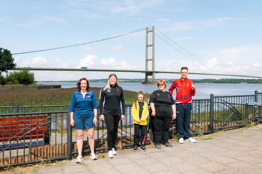 Five people of different ages, all wearing sportswear, stand next to the River Humber, with the Humber Bridge in the background