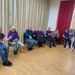 A group of people sat in a circle in a village hall doing exercise with ribbons
