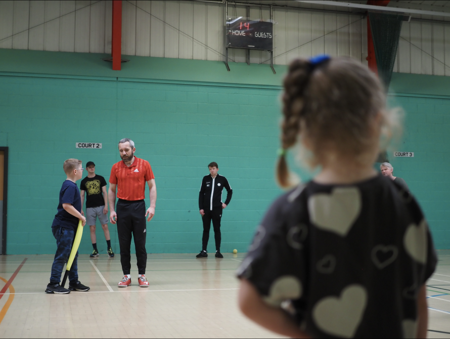 A man in a red t shirt coaching a boy to play blind cricket