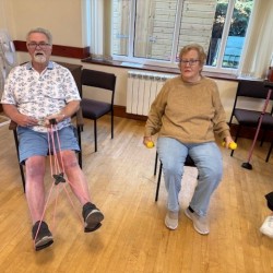 Two people sat on chairs. One is holding dumbbell weight and the other is using resistance bands